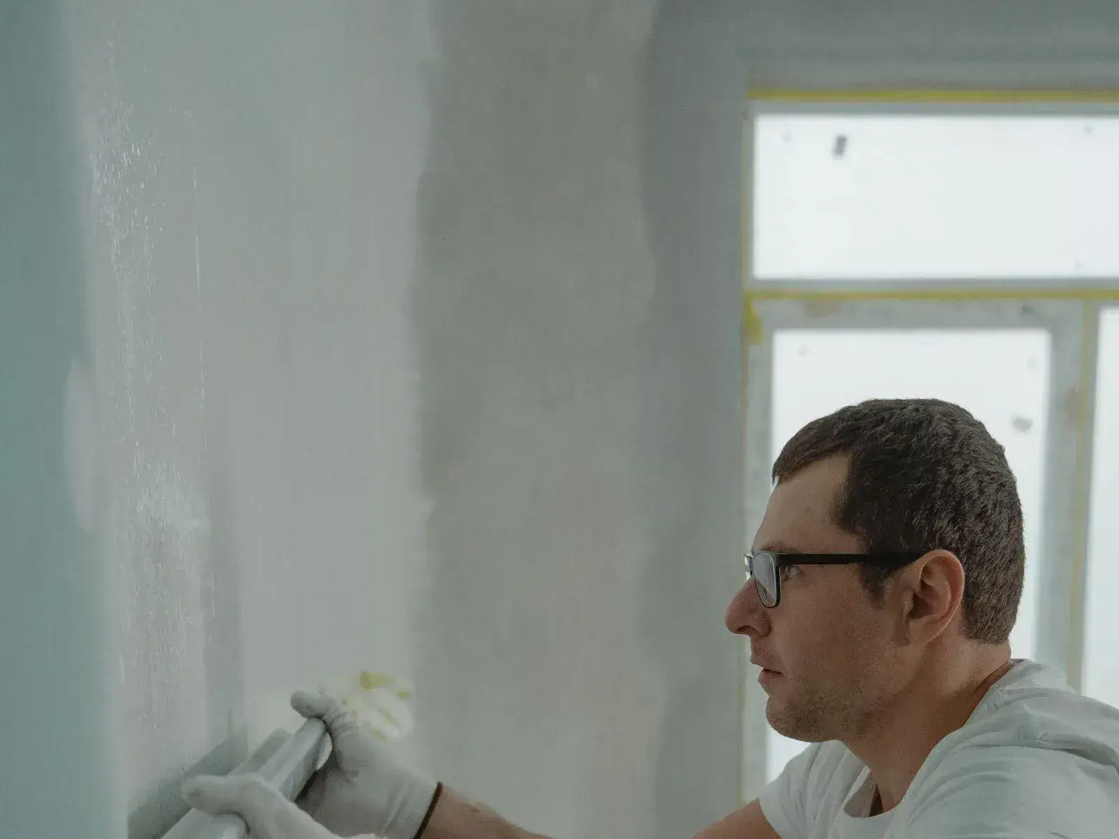 A worker in gloves drawing a jointing knife down a plasterboard wall to fill and smooth the surface.