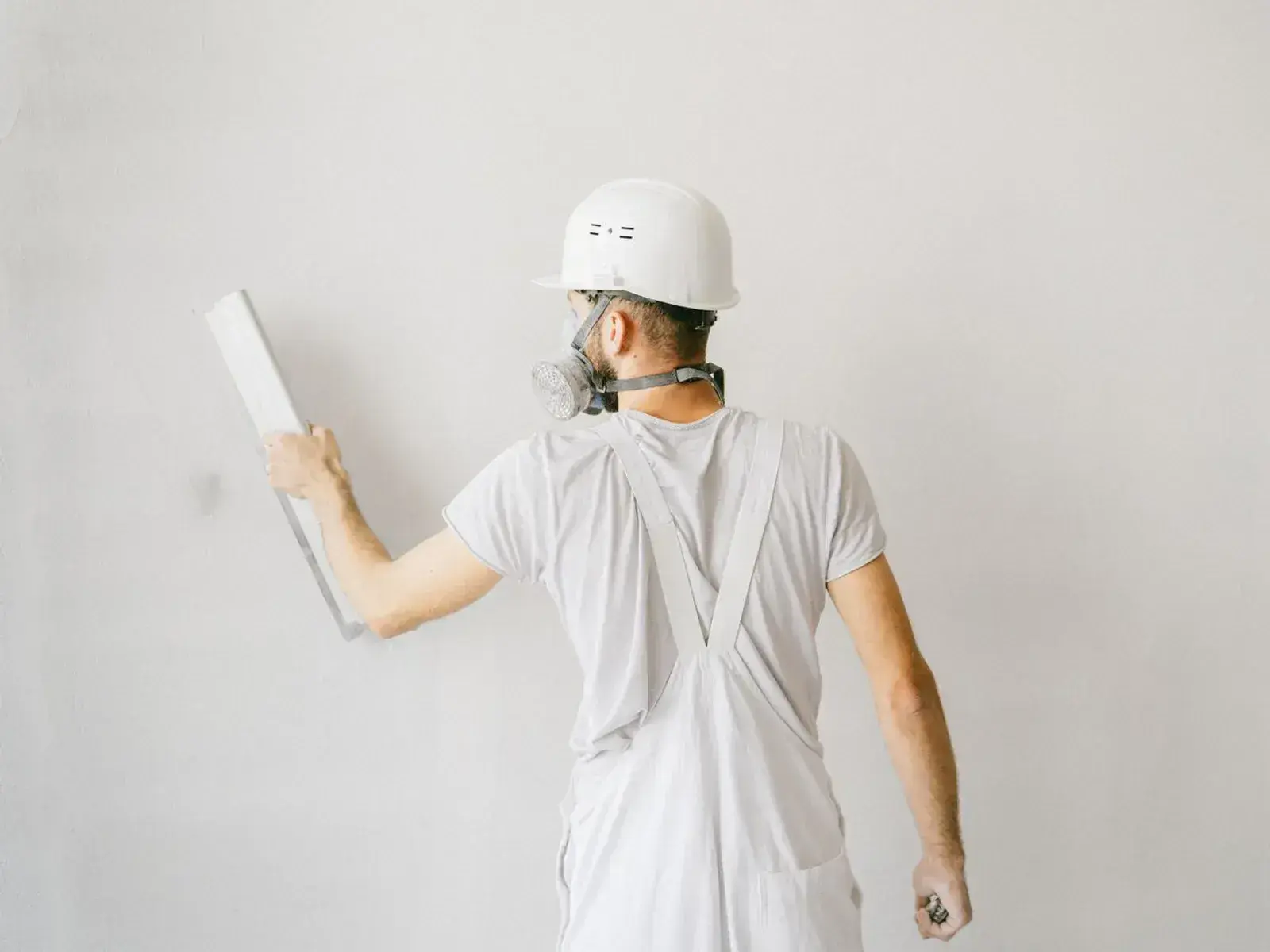A plasterer in white overalls, hard hat and respirator holding a plastering float flat against a smooth white wall.